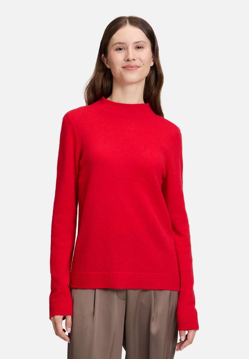 Młoda kobieta z długimi brązowymi włosami, ubrana w czerwoną długą bluzkę i beżowe szerokie spodnie, stoi na tle jednolitego tła.