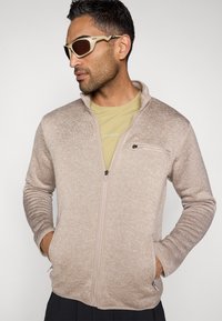 Ανοιχτό καφέ μπουφάν με φερμουάρ και υφή, πλαϊνές τσέπες και τσέπη στο στήθος. Φορεμένο πάνω από ένα κίτρινο πουκάμισο και συνδυασμένο με μπεζ γυαλιά ηλίου.