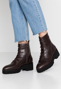 Bottes basses en cuir marron avec une finition texturée, bout rond, design à lacets, détail avec fermeture éclair et semelle extérieure en caoutchouc noire épaisse.