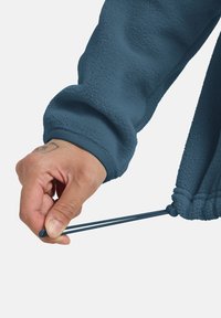 Giacca di fleece blu con polsini a coste. Una mano sta tirando un cordino all'estremità della manica, mettendo in evidenza un elemento di design funzionale.