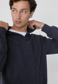 Sudadera con cremallera en un tono azul marino oscuro, con cordones ajustables, mangas largas y un bolsillo tipo canguro. Textura suave y aterciopelada visible.