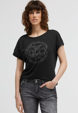 Woman with curly brown hair wearing a black t-shirt with white circular text and faded black jeans, standing with one hand in pocket.