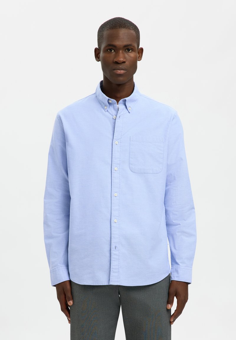 Hombre con camisa azul claro abotonada y pantalones grises de pie sobre un fondo blanco liso con expresión neutral.