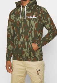 Bluza z kapturem w zielono-brązowych odcieniach moro, z regulowanym sznurkiem przy kapturze i przednią kieszenią typu kangurka. Logo na piersi. Materiał bawełniany.