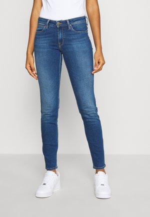 Person, die enge blaue Jeans und weiße Turnschuhe trägt, steht vor einem einfarbigen hellen Hintergrund.