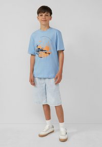 Lichtblauwe kortekout t-shirt met een zwart en oranje strandzonsondergang graphic, gecombineerd met lichtdenim shorts en witte sneakers.