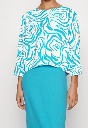 Kobieta ubrana w białą bluzkę z długim rękawem z niebieskimi abstrakcyjnymi zawijasami oraz jednolitą niebieską spódnicę z wysokim stanem.