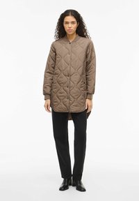 Ruda siuvinėta striukė su apvalia apykakle, priekyje užsegama spaustukais, šoninėmis kišenėmis ir ilgomis rankovėmis, dėvima ant juodų kelnių ir aukštakulnių.