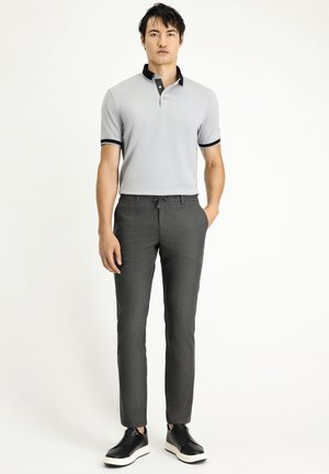 Hellgraues Poloshirt mit schwarzen Kragenakzenten, kombiniert mit dunkelgrauen maßgeschneiderten Hosen und schwarzen Schuhen mit weißen Sohlen. Glatte Textur.