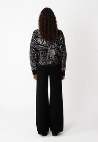 Zwarte trui met abstracte witte geometrische patronen, voorzien van een geribde halslijn en mouwen. Gecombineerd met wijde zwarte broek.