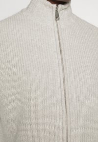 Hellgrauer Strickpullover mit hohem Kragen, Reißverschluss vorne und geripptem Muster. Verfügt über ein weiches, voluminöses Maschenmuster.