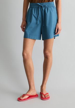 Person trägt blaue Shorts mit Kordelzug und rote Flip-Flops und steht auf einem schlichten grauen Boden vor einem hellgrauen Hintergrund.