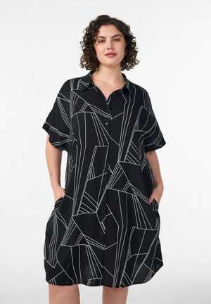 Kvinde med krøllet hår iført en sort kjole med knapper og hvide geometriske linjer, stående med hænderne i lommerne mod en hvid baggrund.