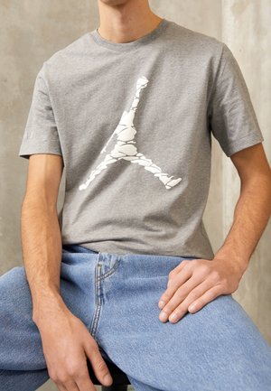 Grå bomulds T-shirt med et stort hvidt grafisk motiv, der ligner et logo. Korte ærmer, rund halsudskæring, båret med lyseblå jeans.