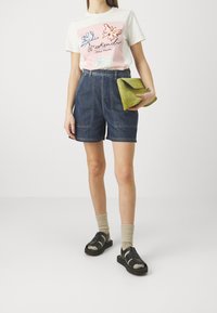 Camiseta blanca con gráficos de mariposas en rosa y azul, combinada con pantalones cortos de mezclilla de tiro alto. Un bolso verde, calcetines grises y sandalias negras completan el look.