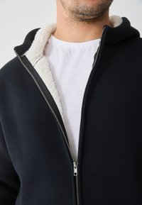 Černá mikina na zip s bílým sherpa podšívkou, s lesklým zipem, navrstvená přes bílý tričko. Hladká textilie.