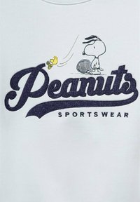 Svetlo modra majica s kapuco, ki vsebuje temno morsko modro vezeno logotip "Peanuts", s kartonskimi figuricami Snoopyja in Woodstoka, ki igrata tenis.