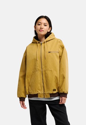 Woman with long dark hair wears a loose yellow hooded jacket with front pockets and black pants, standing against a plain light background.