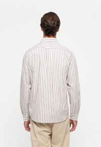 Man with wavy brown hair wearing a white long-sleeve shirt with vertical red stripes and beige pants, facing away on a white background.