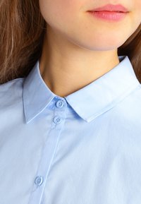 Světle modrá košile s klasickým límcem, která má dva knoflíky u krku a hladkou, strukturovanou látku.