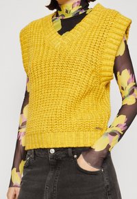 Geel gebreid mouwloos sweater met een V-hals en ribgeborduurde randen, met een textuurpatroon en gecombineerd met een bloemen mesh-top.