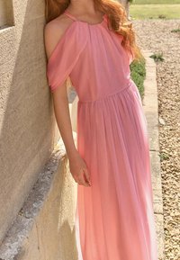 Robe rose longue jusqu'au sol avec un design plissé, manches épaules dénudées, tissu léger et taille froncée. Légère brillance et texture douce.