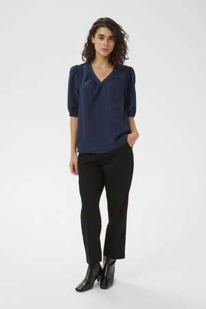 Mujer con cabello rizado que lleva una blusa azul marino de cuello en V, pantalones cortos negros y botines negros, de pie con una mano en el bolsillo.