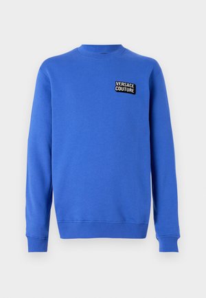 Sweatshirt azul de manga longa com decote redondo, apresentando punhos e barra canelados, com um pequeno patch preto e branco "Versace Couture" no peito.