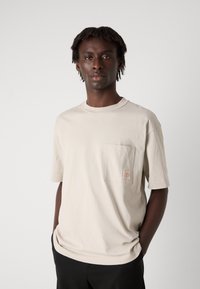 Šviesiai rudas medvilninio t-shirt marškinėliai su trumpomis rankovėmis, apvaliu kaklu ir kairiajame krūtinės pusėje esančia kišenėle su maža logotipų etiketė. Paprastas, klasikinis dizainas.