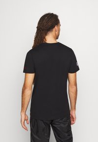 T-shirt en coton noir avec un col rond, des manches courtes et un petit logo sur la manche gauche. Texture lisse et coupe décontractée.