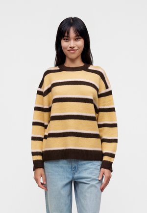 Jeune femme aux cheveux foncés portant un pull rayé jaune, marron et beige et un jean bleu clair, debout devant un fond clair uni.