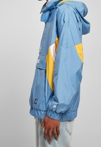 Giacca a vento leggera di colore blu chiaro con accenti gialli e bianchi, polsini elasticizzati e tasca sul petto. Realizzata in materiale liscio e leggero.