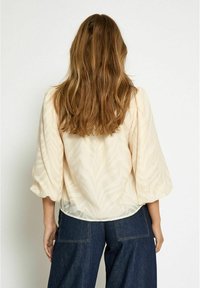 Peach-colored blouse with a semi-sheer fabric, featuring a textured leaf pattern, puffed sleeves, and a loose fit paired with dark denim pants.