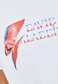 Hvid bomulds t-shirt med et grafik i rød og blå nuancer, tekst der siger "DAVID BOWIE ALAIDDI", og en detaljeret ansigtsillustration.
