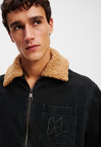 Chaqueta de mezclilla negra con cuello de sherpa color tostado, cierre de cremallera y un bolsillo con parche que presenta un detalle de logotipo bordado. Tejido texturizado.