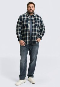 Flanellen overhemd in marineblauw, groen en wit ruitpatroon met twee borstzakken; gecombineerd met een grijs T-shirt en blauwe jeans, afgewerkt met witte sneakers.