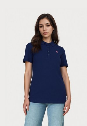 Jeune femme aux longs cheveux foncés portant un polo bleu marine avec des boutons et un jean bleu clair, debout devant un fond blanc uni.