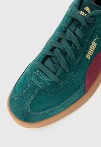 Sneaker Puma din suede verde cu șireturi verzi închis, o dungă laterală maron, logo-ul aurie Puma și talpă din cauciuc, pe fundal alb.
