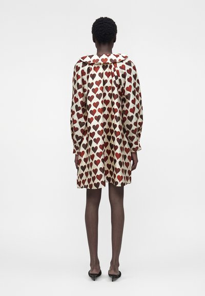 Γυναίκα που στέκεται με την πλάτη, φοράει κρεμ φόρεμα με κόκκινο σχέδιο καρδιάς, μακριά μανίκια και μαύρα mule με τακούνι.