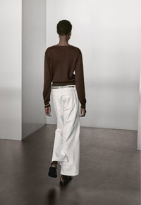 Maglione lavorato a maglia marrone abbinato a pantaloni bianchi larghi. L'outfit presenta una cintura metallica e scarpe nere, visto da dietro.