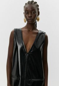 Must musta, ilma varrukat, läikivast materjalist kleit, millel on sügav V-avasse ja eesosal taskud. Omab puhast disaini ja sujuvat tekstuuri.