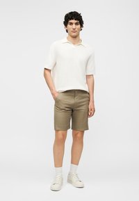 Ένας νεαρός άνδρας στέκεται με το ένα χέρι στην τσέπη, φοράει ένα λευκό πλεκτό πόλο με κοντά μανίκια, χακί σορτς, λευκές κάλτσες και λευκά αθλητικά παπούτσια.