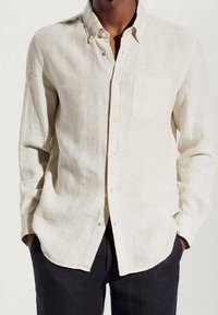 Uomo che indossa una camicia di lino beige a maniche lunghe e bottoni, con una tasca sul petto e pantaloni scuri, con le mani in tasca.