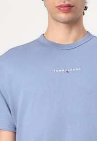 Šviesiai mėlynas medvilninis marškinėlis su apvalia iškirpte, su balta siuvinėta "TOMMY JEANS" logotipu krūtinėje. Trumpomis rankovėmis, tekstūruotas audinys.