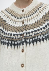 Gros plan sur un cardigan en maille crème avec des boutons ronds foncés et un motif nordique marron, noir et blanc sur la poitrine et les épaules.