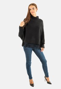 Pull en maille noir avec un col haut, doté d'un ourlet à franges. Associé à un jean bleu marine et des talons noirs. Texture lisse.