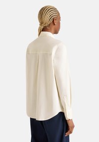Camicia color crema con piega sulla schiena, maniche lunghe e polsini con bottoni. Realizzata in tessuto morbido con una vestibilità rilassata e orlo arrotondato.