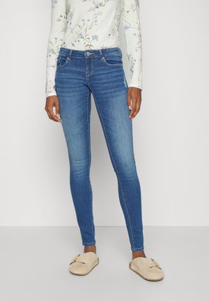 Jeans ajustados azules con un diseño desgastado, que incluyen bolsillos frontales y un cierre de botón metálico, combinados con zapatos beige tipo slip-on.