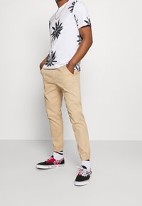 Witte t-shirt met zwarte bloemenprints, beige tapered broek en zwarte sneakers met roze accenten en een geruit patroon. Casual pasvorm.