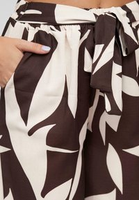 Pantalons en tissu à motifs marron et crème avec une taille élastique, des poches latérales et une ceinture à nouer, présentant de grandes formes de feuilles dans des couleurs contrastantes.